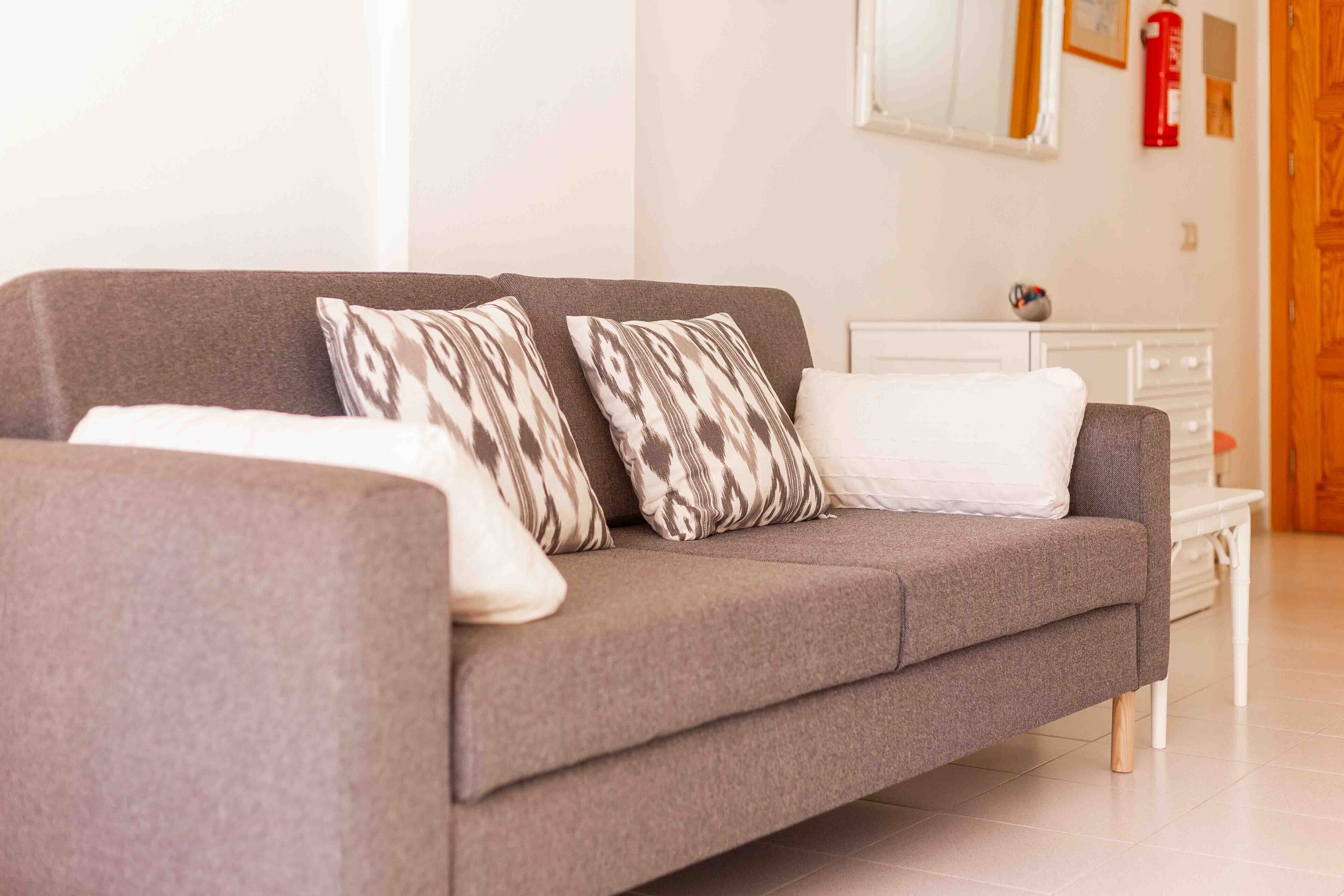Ein modernes Wohnzimmer mit einem grauen Sofa, gemusterten Kissen, einem weißen Beistelltisch und einer weißen Kommode.