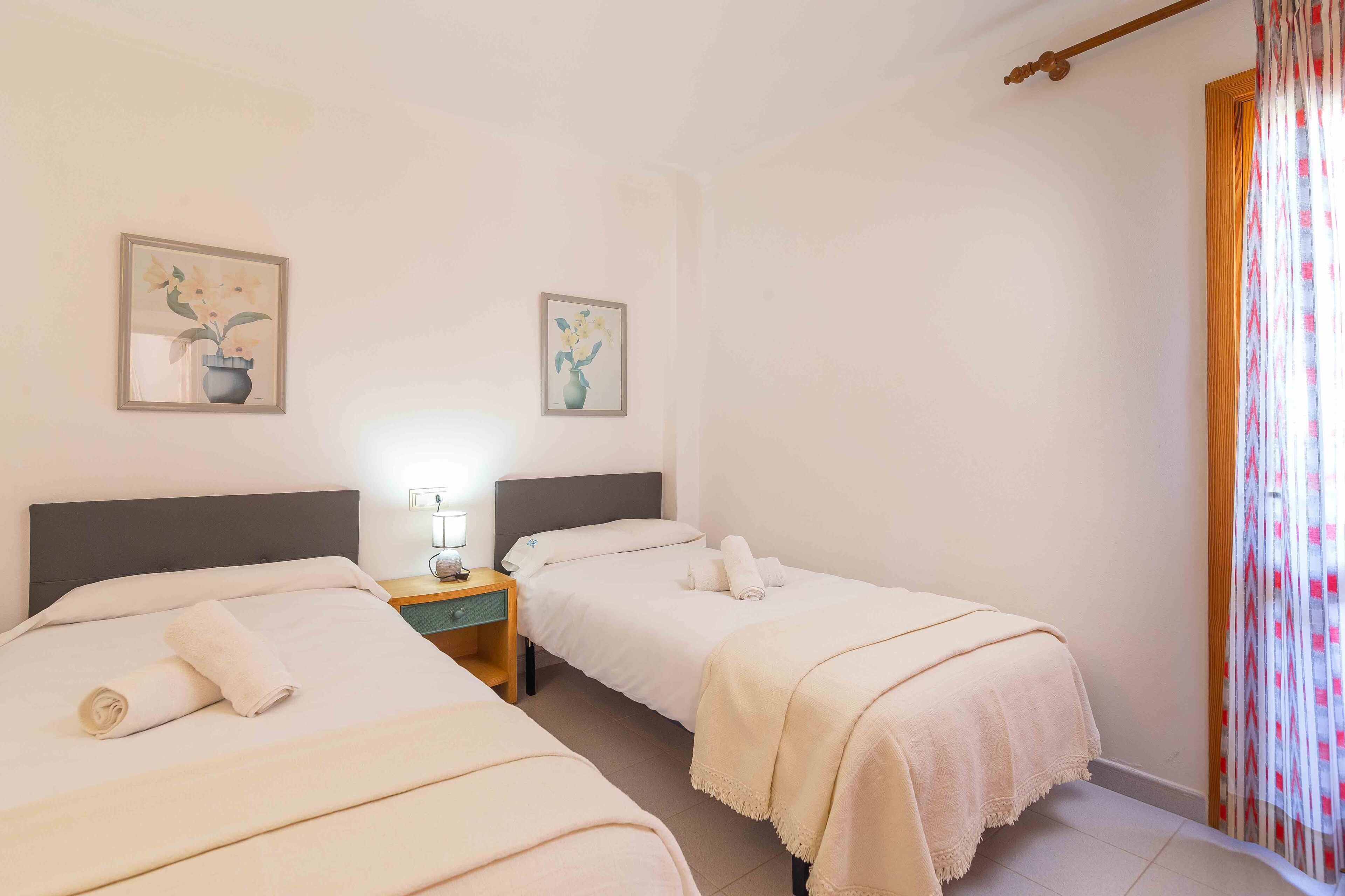 A hotel room with two beds, a nightstand with a lamp, and two framed floral paintings on the wall.