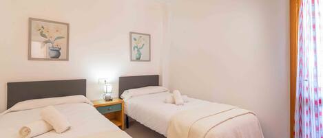 A hotel room with two beds, a nightstand with a lamp, and two framed floral paintings on the wall.