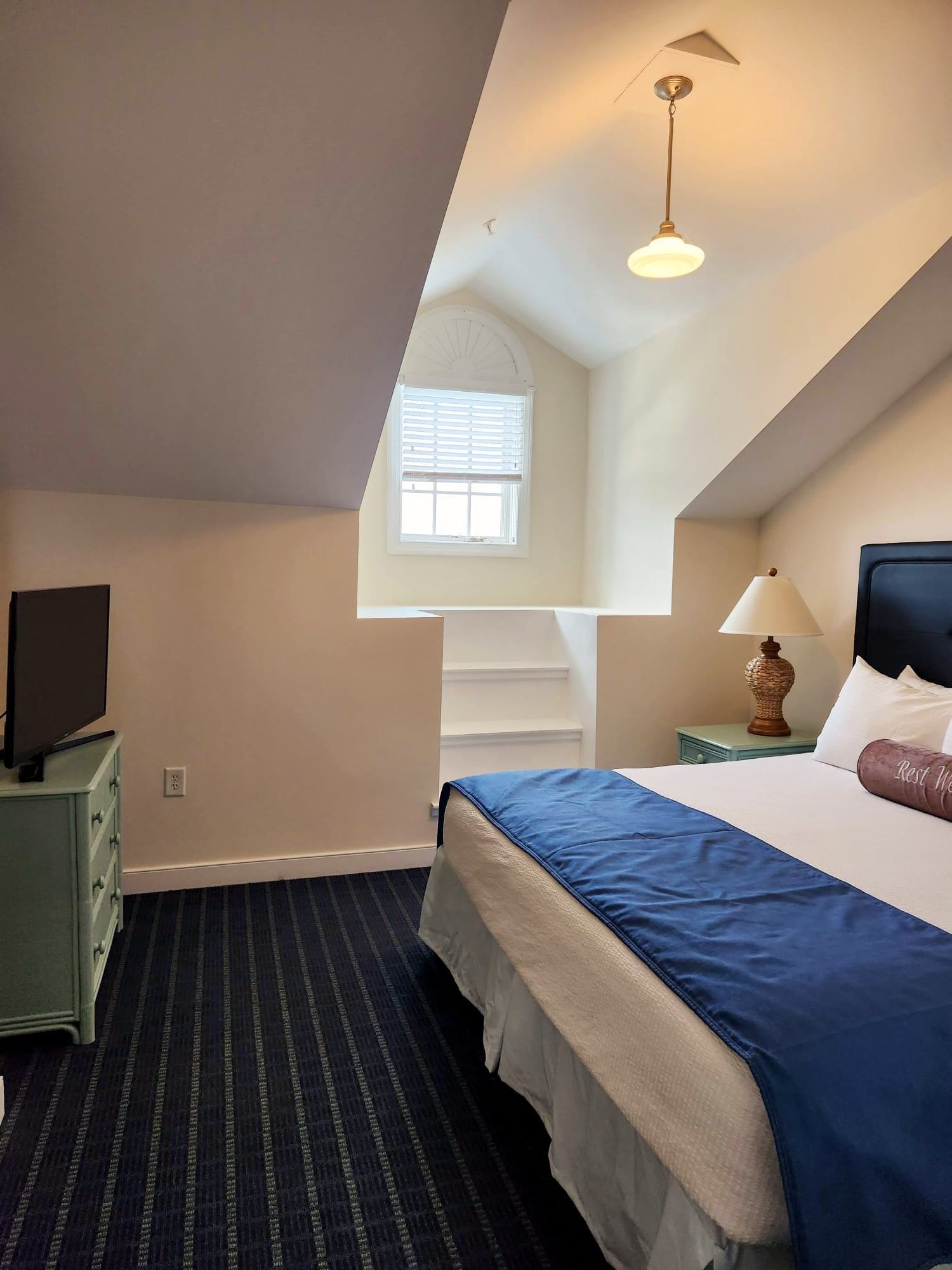 A hotel room with a bed, a desk with a lamp, a television, and a window with blinds.