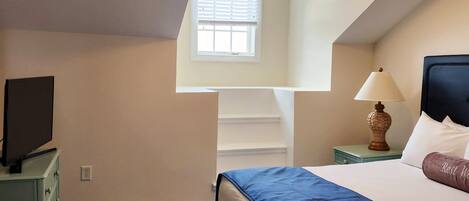 A hotel room with a bed, a desk with a lamp, a television, and a window with blinds.