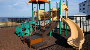 A playground with a slide and climbing structures, surrounded by a fence and a building in the background. - Waterside Resort (Roper)