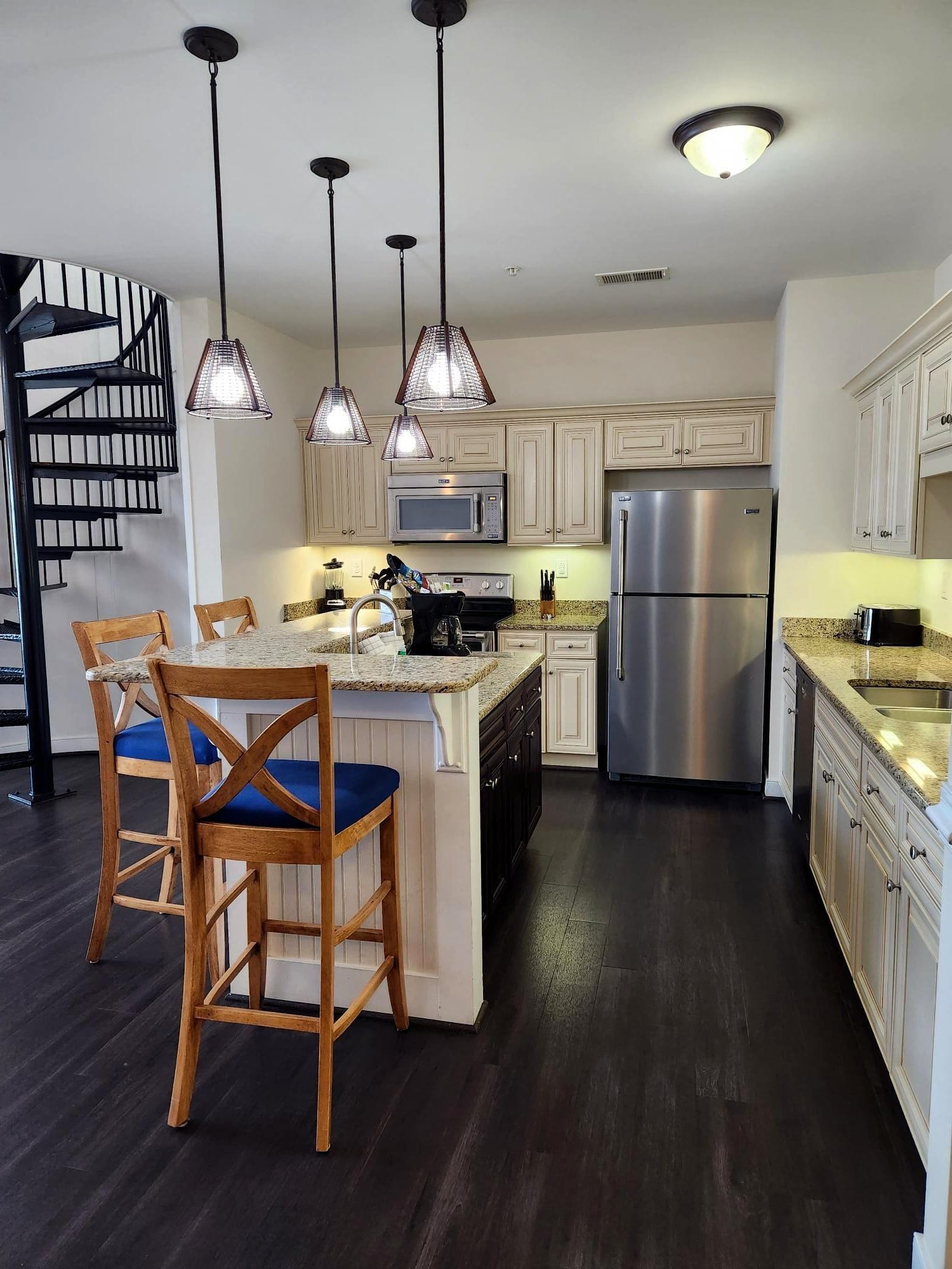 A modern kitchen with a central island, stainless steel appliances, pendant lighting, and a dark wood floor.