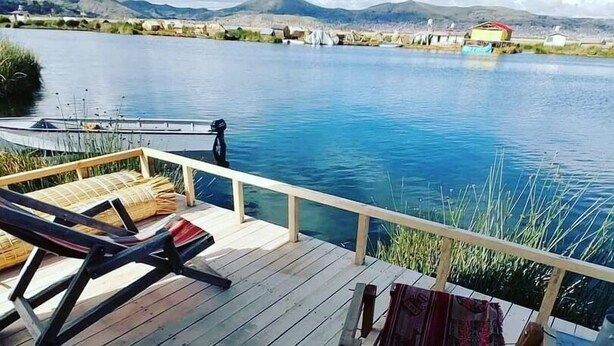 Une terrasse en bois avec des chaises longues et une table, d’où l’on peut admirer un lac paisible avec des montagnes en arrière-plan.