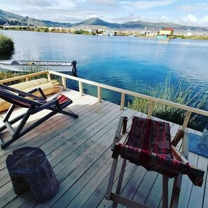 Een houten terras met lounge stoelen en een tafel, met uitzicht op een rustig meer met bergen op de achtergrond.
