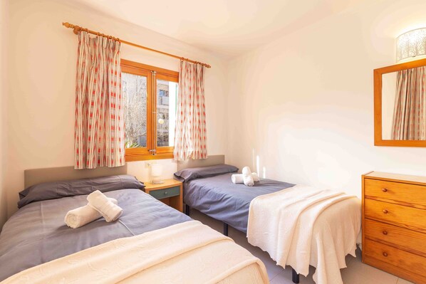 Una habitación con dos camas individuales, un aparador de madera, una mesita de noche y una ventana con cortinas.
