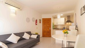 A modern living room with a grey sofa, a white dining table, and a kitchen area with cabinets and a microwave.
