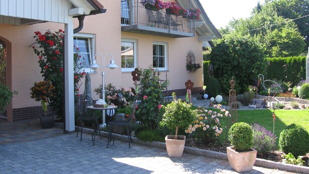 A two-story building with a balcony adorned with flowers, a small outdoor seating area with a table and chairs, and a well-maintained garden with various plants and a lawn. - Family-friendly apartment near Legoland for 5 pers.                (Kammeltal)