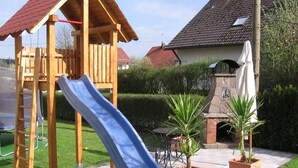 A wooden play structure with a slide, a brick fireplace, and potted plants in a garden area. - Family-friendly apartment near Legoland for 5 pers.                (Kammeltal)