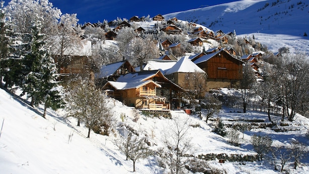 A snow-covered mountain village with wooden cabins and a ski slope in the background.