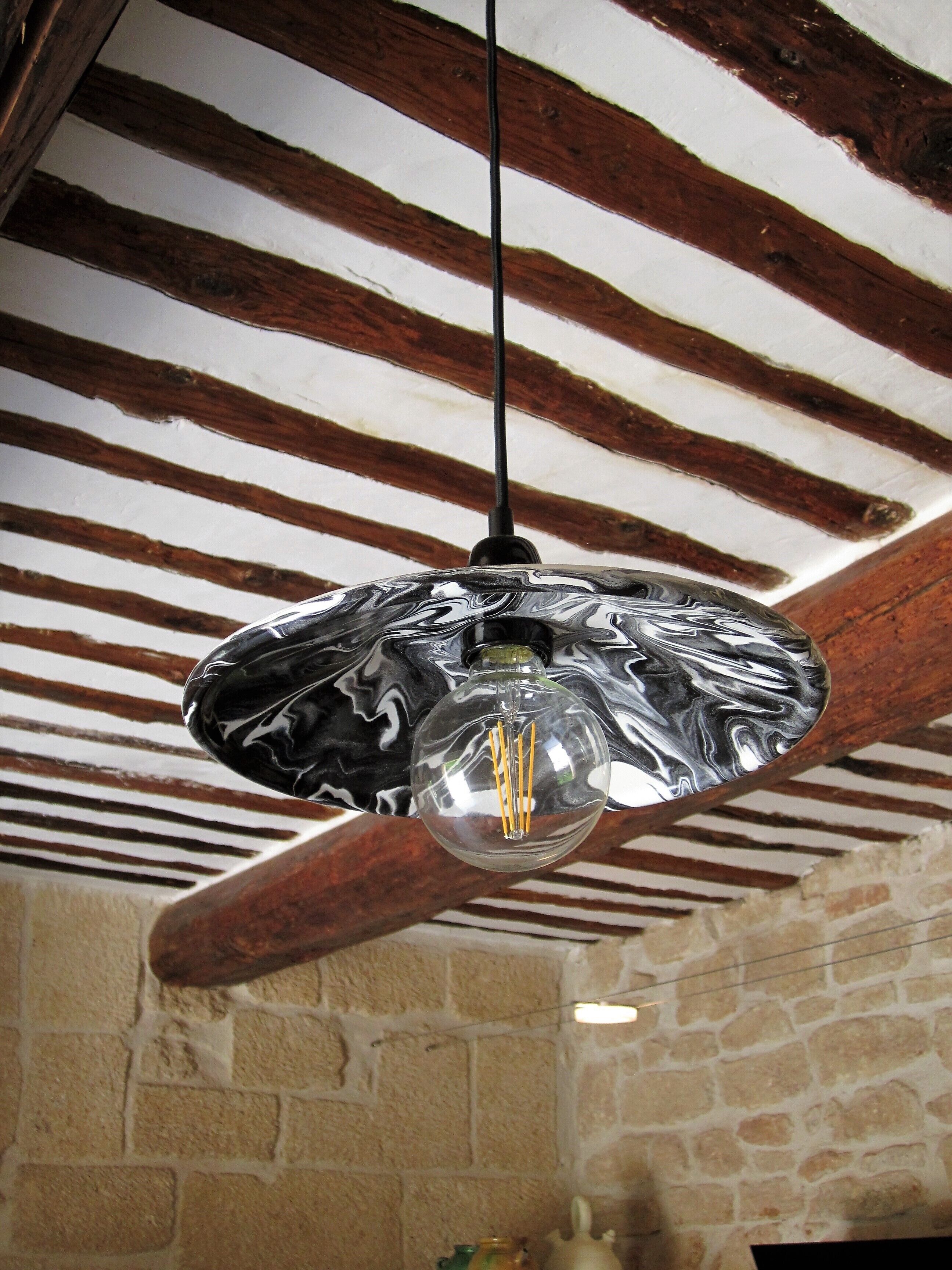 Un luminaire suspendu, de type suspension, avec un revêtement métallique réfléchissant, dans une pièce dotée de poutres apparentes en bois et de murs en pierre.