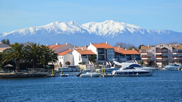 A marina with boats docked, a mountain range in the background, and buildings with red-tiled roofs. - La Marina - Charming Holiday Home in Residence with Swimming Pool (Saint-Cyprien)
