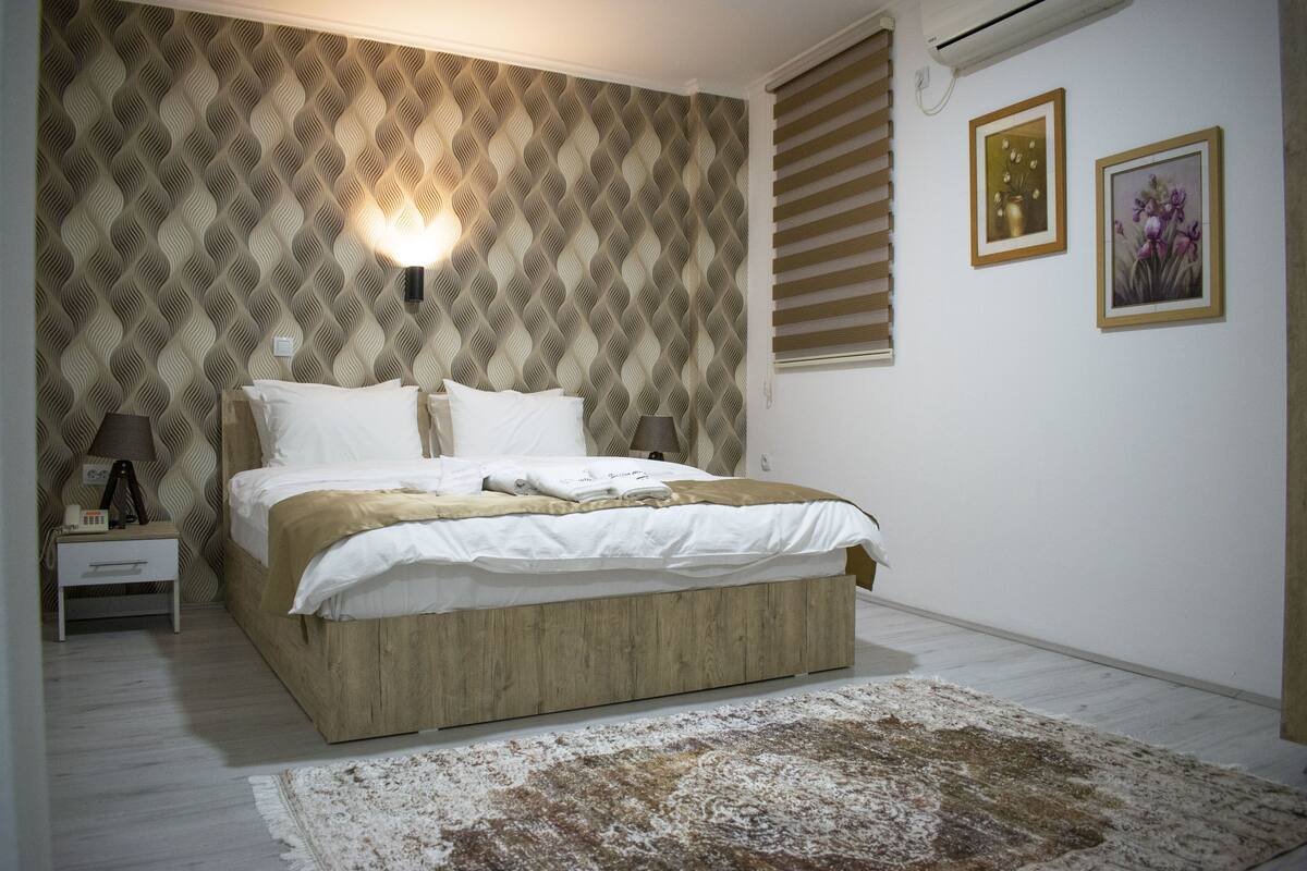 A bedroom with a patterned wallpaper, a bed with white bedding, a nightstand with a lamp, and two framed pictures on the wall.
