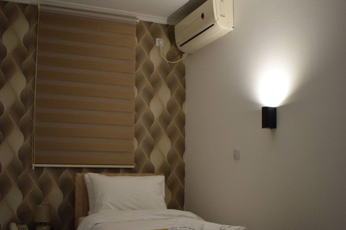 A bedroom with a bed, a nightstand, a wall-mounted air conditioner, and a wall-mounted light fixture.
