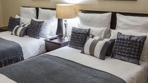 Habitación de hotel con dos camas, cada una con ropa de cama blanca y mantas grises. Entre las camas, un mesita de noche con una lámpara.