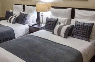 Habitación de hotel con dos camas, cada una con ropa de cama blanca y mantas grises. Entre las camas, un mesita de noche con una lámpara.
