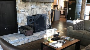 Ein gemütliches Wohnzimmer mit Kamin aus Stein, Ledersesseln, einem Couchtisch und einer Küche im Hintergrund.