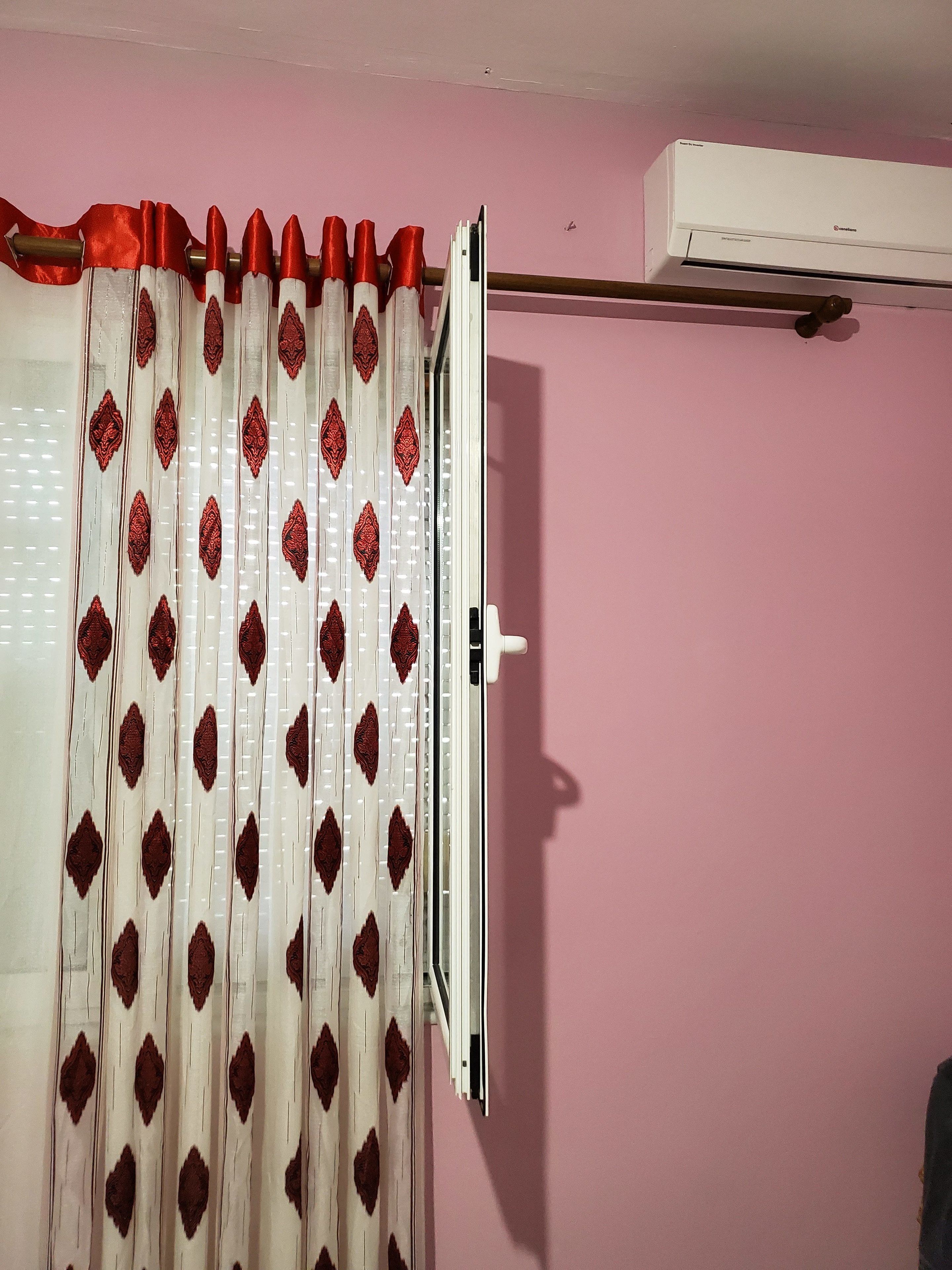 A room with a pink wall, a window with a white curtain featuring a diamond pattern, and a white air conditioning unit mounted on the wall.