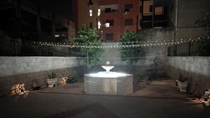 Une cour intérieure avec une fontaine centrale, entourée de plantes en pot et illuminée de guirlandes lumineuses.