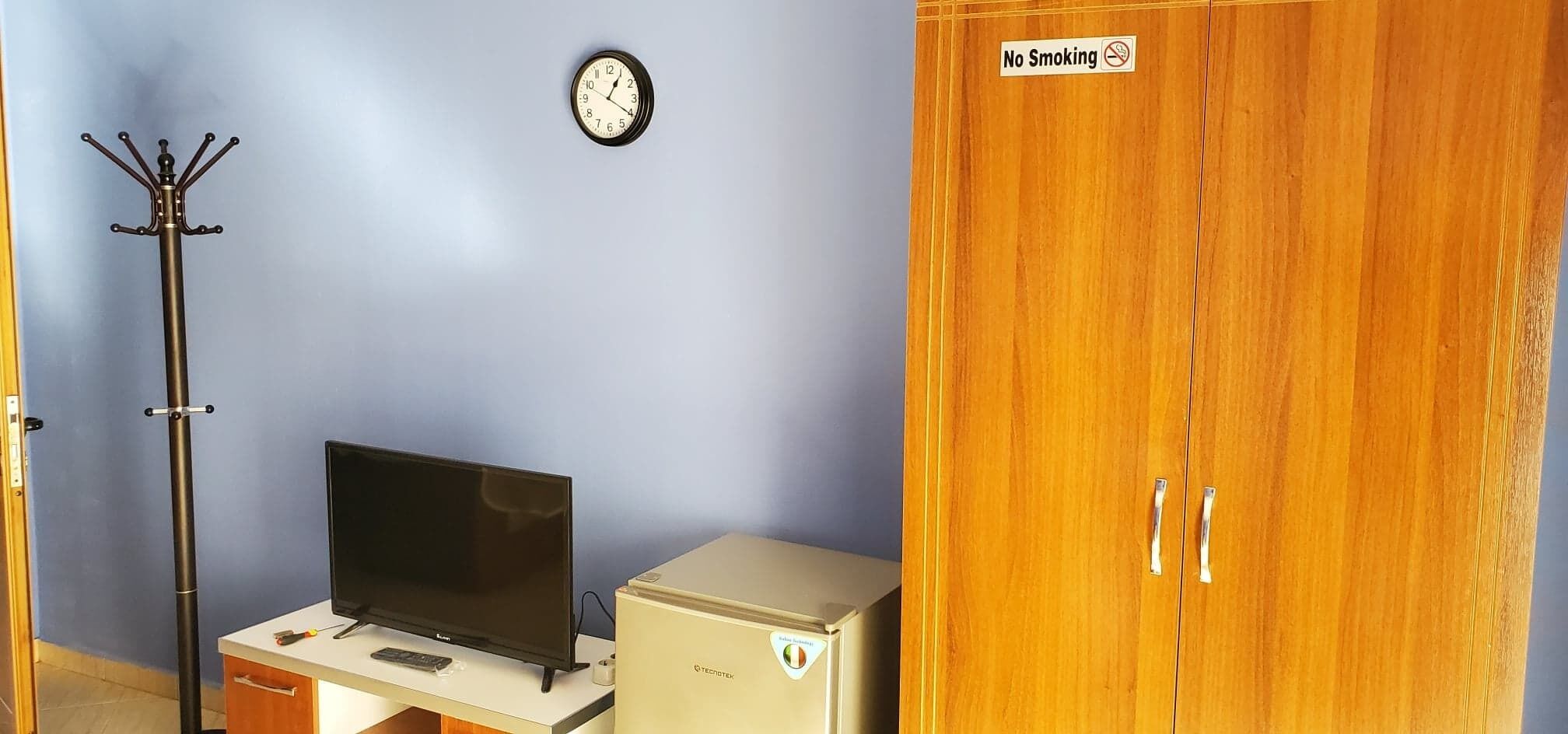 A room with a television, a clock, a coat rack, a cabinet, and a wardrobe with a 'No Smoking' sign.