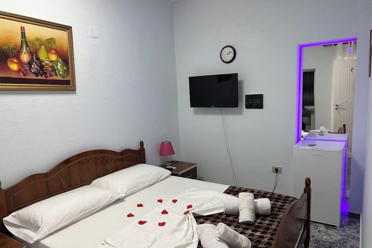 A hotel room with a bed, a television, a clock, a mirror, and a small table with a lamp.