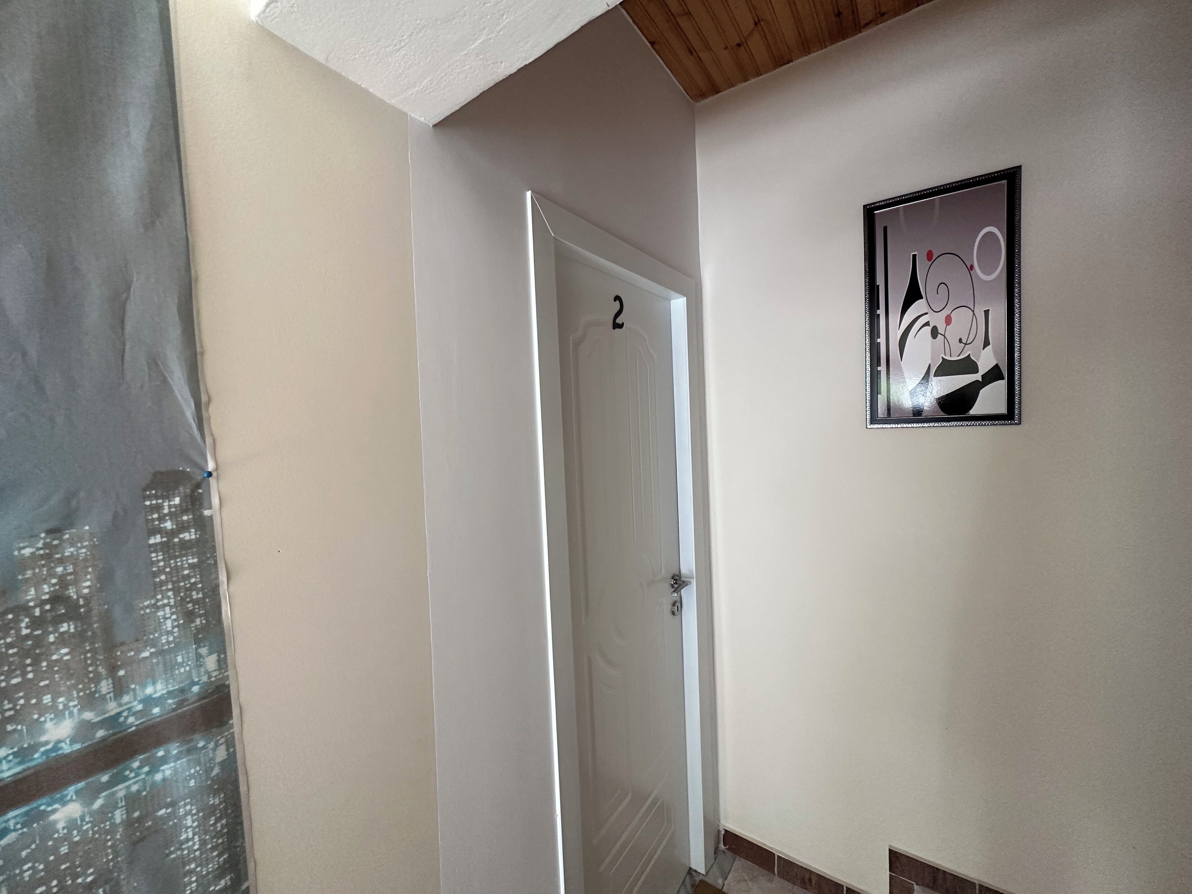 Um corredor com uma porta branca numerada 2, uma obra de arte abstrata emoldurada na parede e uma janela parcialmente visível com vista para a cidade.