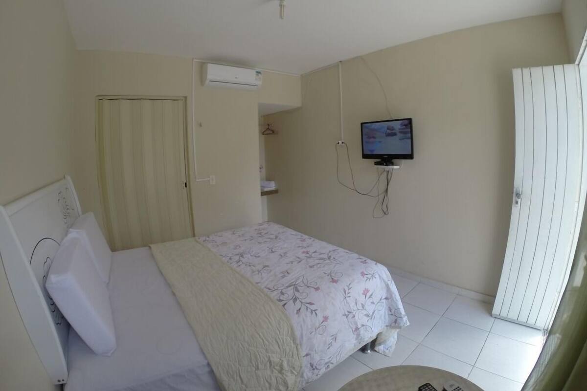 A bedroom with a bed, a television mounted on the wall, a ceiling fan, and a window with blinds.