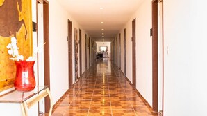 Un largo y estrecho pasillo de hotel con suelo de baldosas, puertas de madera y un jarrón rojo con flores blancas sobre una mesa.