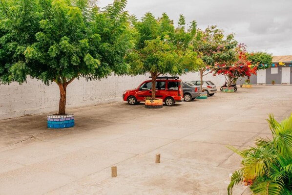 Un aparcamiento con un coche rojo, un coche gris y una motocicleta. Se ven árboles y una pared blanca al fondo.