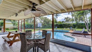 Eine überdachte Terrasse mit einem Glastisch und Korbstühlen, mit Blick auf einen Swimmingpool und einen Basketballplatz.