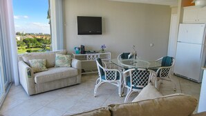Ein Wohnzimmer mit einer Couch, einem Glastisch mit Stühlen, einem wandmontierten Flachbildfernseher und Blick ins Freie.