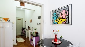 A dining area with a round table, white chairs, a red vase with flowers, and a colorful abstract painting on the wall. - Omar do Rio - NSC1001 (Rio de Janeiro)