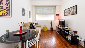 A compact living space with a round dining table, a sofa, a television, and a window with blinds. - Omar do Rio - NSC1001 (Rio de Janeiro)