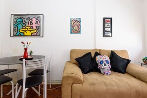 A living room with a tan sofa, black cushions, a red vase with flowers, and a skull-patterned pillow. There are framed artworks on the wall and a small table with a red vase and a cactus.