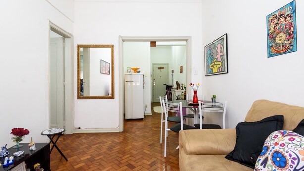 A living room with a herringbone floor, a beige sofa, a small table with a vase, and a mirror with a gold frame. - Omar do Rio - NSC1001 (Rio de Janeiro)