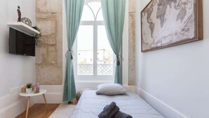 A bedroom with a bed, a small table with a plant, a wall-mounted TV, and a window with curtains.