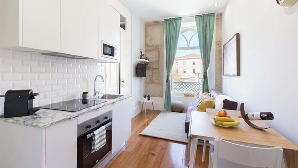 A compact kitchen with white cabinets, a microwave, and a coffee machine. There's a small table with a bowl of fruit and a camera. A window with green curtains and a view of buildings is visible.