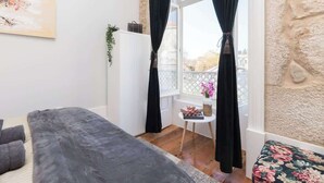 A cozy room with a bed, a small table with a vase of flowers, and a view of the outside through a window with curtains.