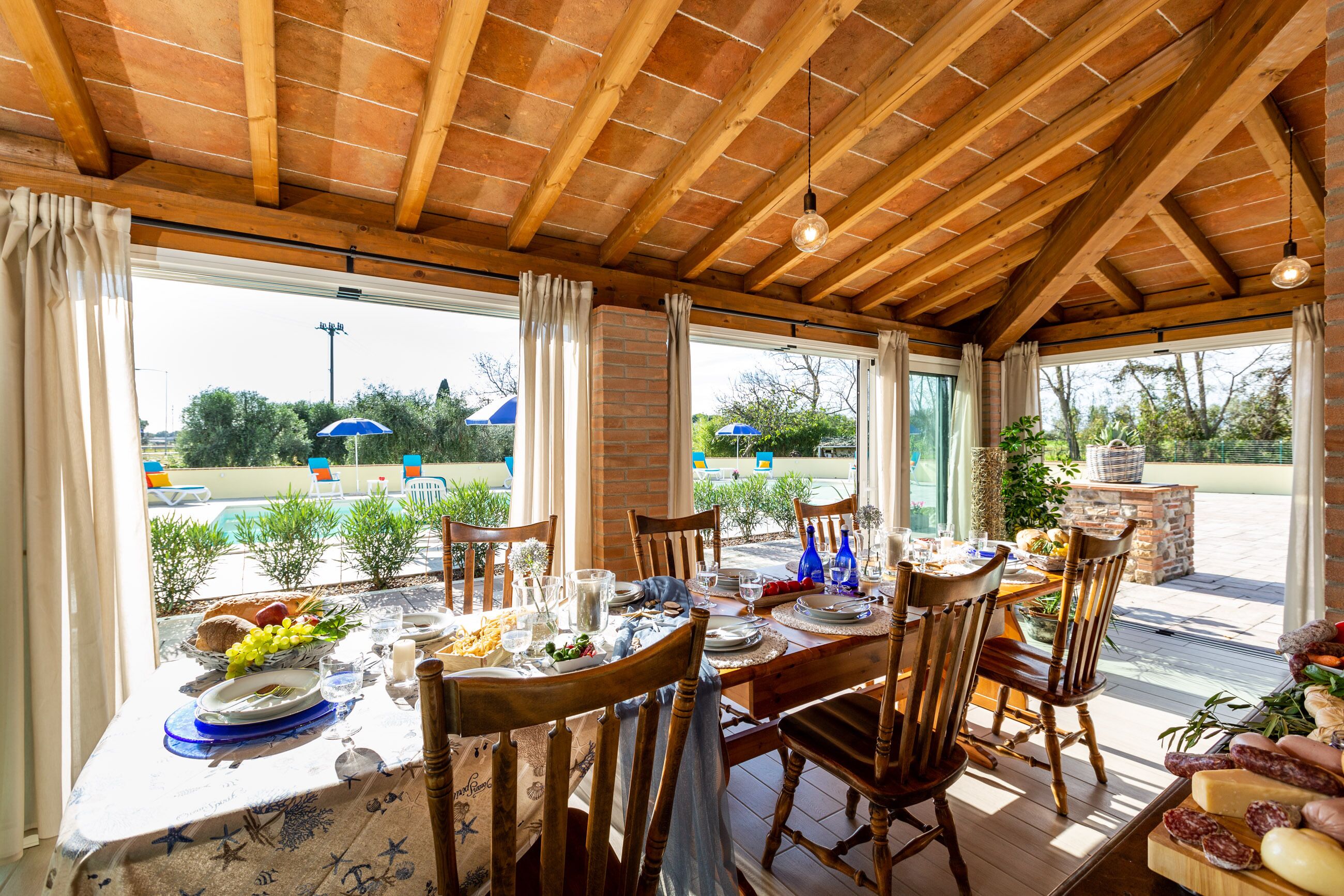 A dining area with a wooden ceiling and large windows overlooking a pool and garden.