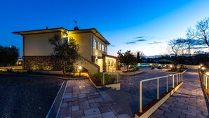 A two-story building with a stone facade and a flat roof, surrounded by a paved walkway and landscaped areas. - Il Nocciolo 12, Emma Villas (Cecina)