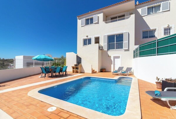 A rooftop swimming pool with a clear blue pool, surrounded by a tiled patio, and a two-story building with white walls and shutters.
