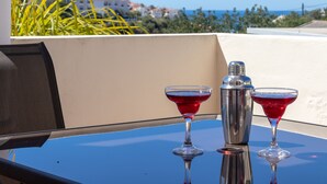 Uma mesa com dois copos de martini e um coqueteleira em uma varanda com vista para edifícios e para o mar.
