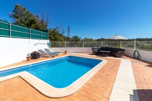 Ein rechteckiger Swimmingpool mit weißem Rand, umgeben von einer Terrasse mit Fliesenboden. Liegestühle und ein Tisch mit einem grünen Sonnenschirm stehen bereit. Ein weißer Zaun begrenzt das Gebiet, und im Hintergrund ist eine grüne Wand zu sehen.