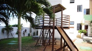 Un terrain de jeux avec un toboggan et des structures d’escalade, entouré d’un bâtiment blanc et d’un palmier.