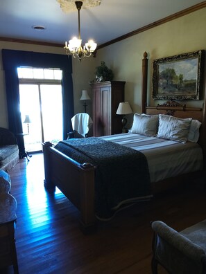 A hotel room with a large bed, a wooden wardrobe, a desk, and a chair. There is a chandelier hanging from the ceiling and a painting on the wall.