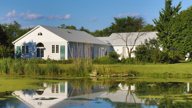 A white building with a gabled roof, surrounded by a pond and greenery. - Inntiquity A Country Inn (Logansport)