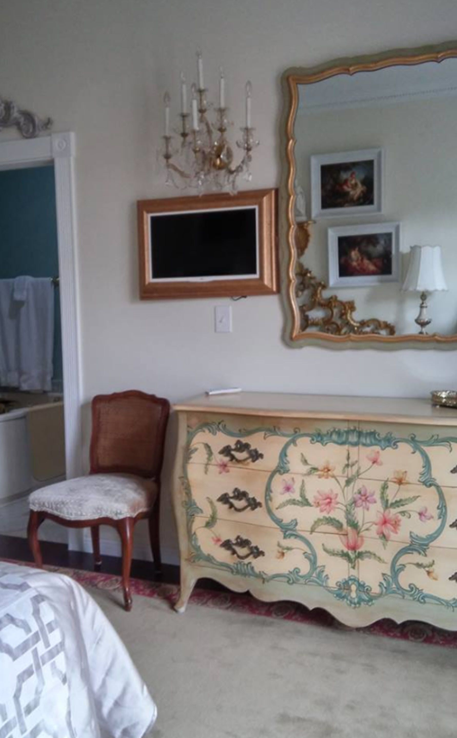 A bedroom with a wooden chair, a dresser with floral patterns, a mirror with an ornate frame, and a chandelier.
