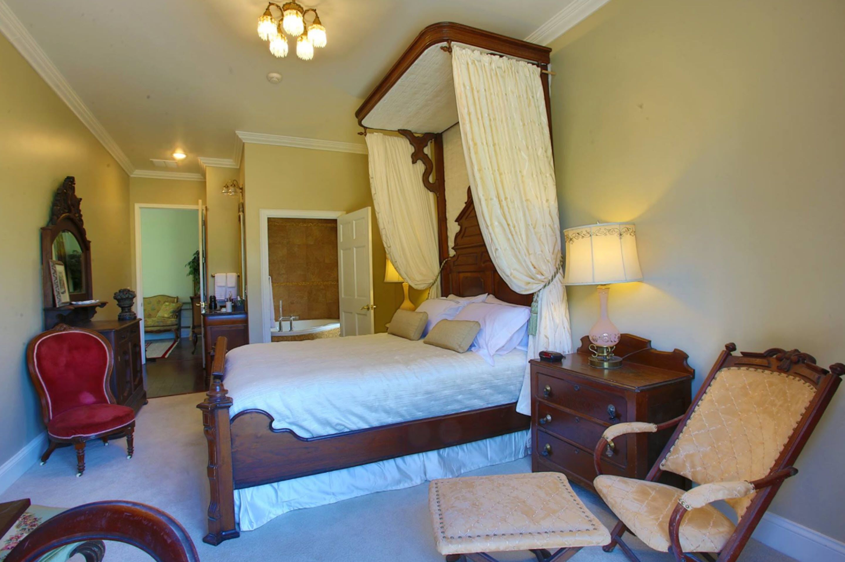 A four-poster canopy bed with a wooden frame and white bedding, a red upholstered chair, a wooden nightstand with a lamp, and a mirror on the wall.