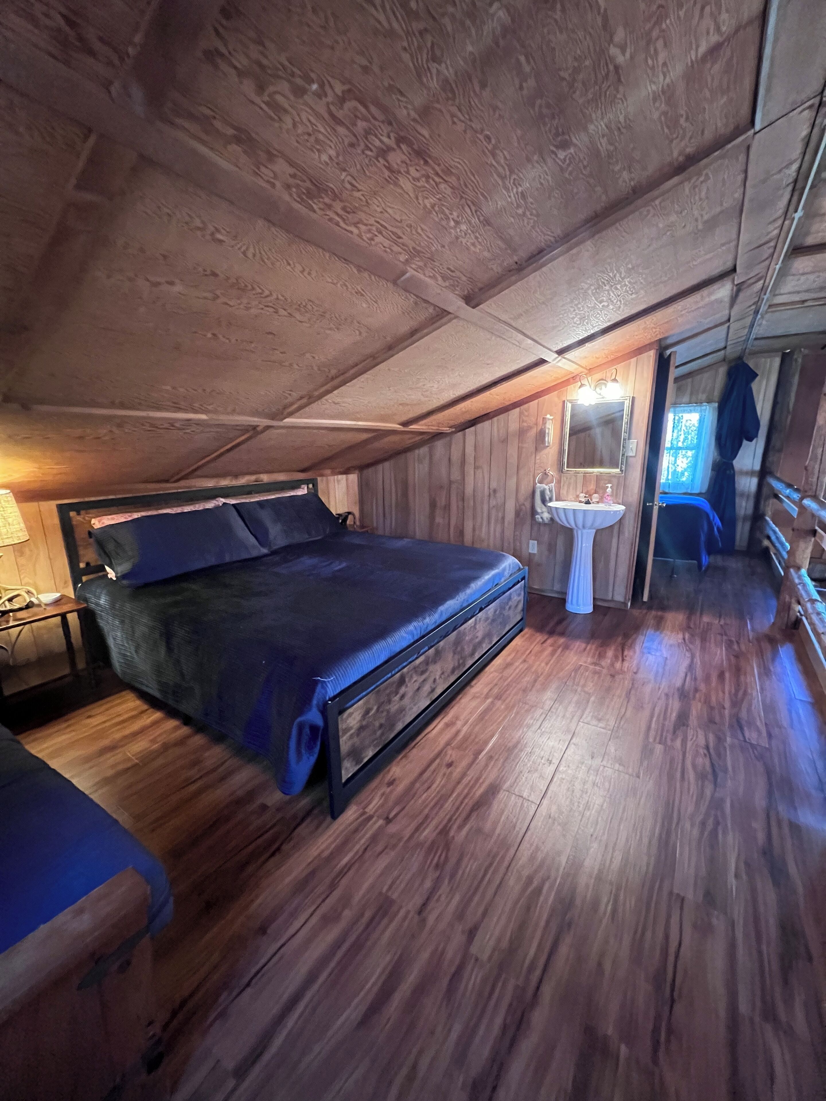 A wooden cabin bedroom with a bed, a bedside table, a mirror, and a window.