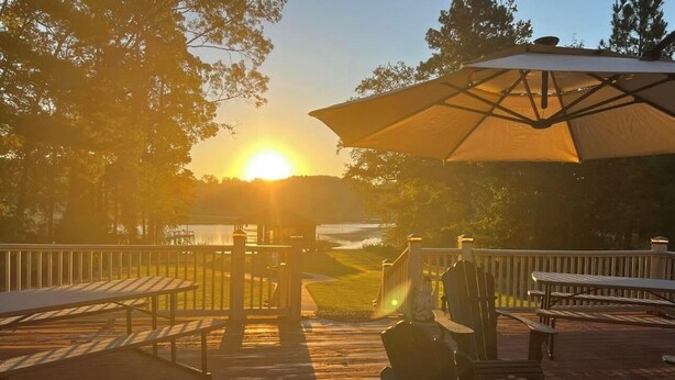 A wooden deck with picnic tables and chairs, a large umbrella, and a view of the sunset over a body of water. - Waterfront Retreat in Toledo Bend (Many)